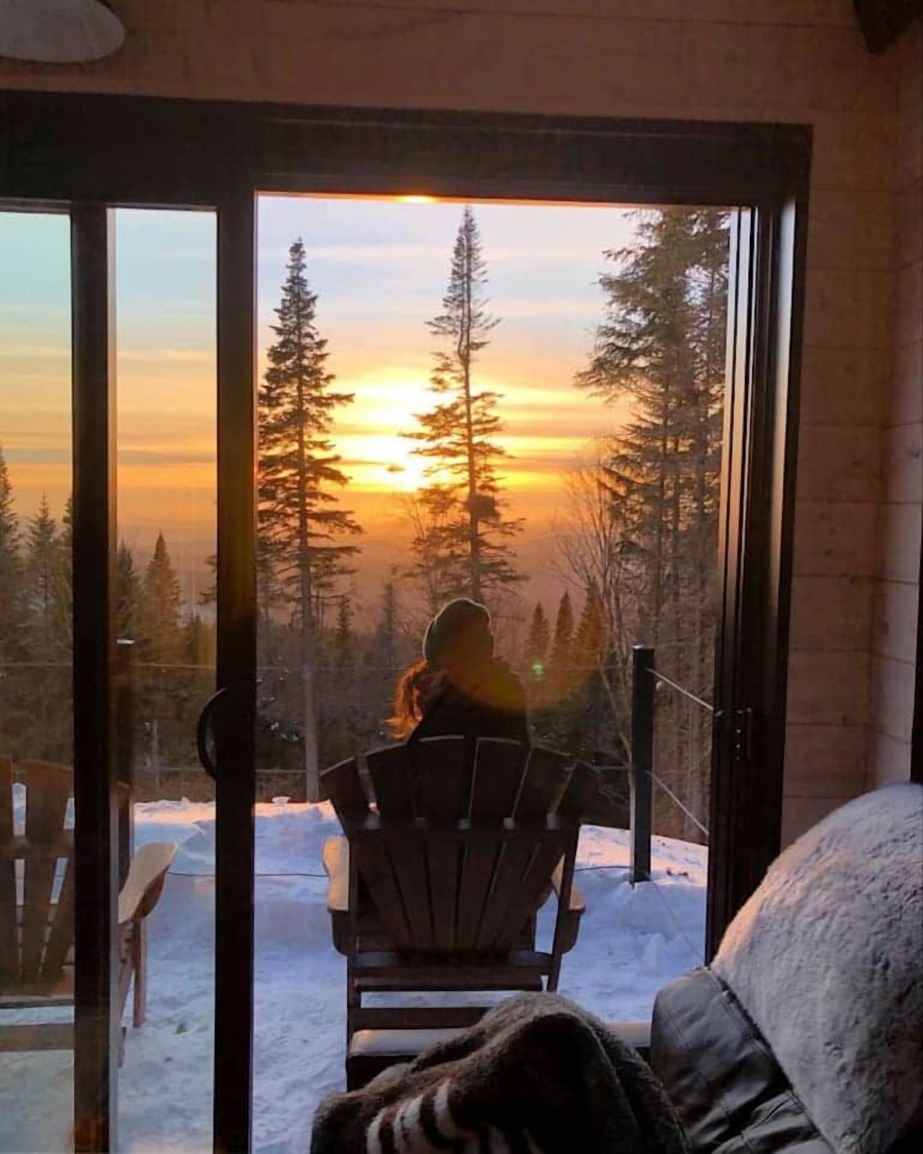 En person sitter på en stol framför en glasdörr med utsikt över ett snötäckt landskap och träd under solnedgången.