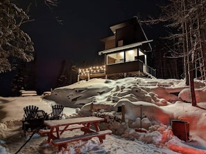Uma área externa coberta de neve, com um deck de madeira, uma mesa de piquenique e cadeiras.