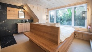 A bedroom with a wooden bed, a built-in bench, a window with a view of trees, and a kitchen area with a sink and cabinets. - MAO / Micro-Chalet / Private Thermal Experience near Quebec (Capitale-Nationale)