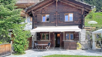 Un chalet en bois avec une façade en pierre, un patio couvert avec une table et des chaises, et une vue sur la verdure environnante.