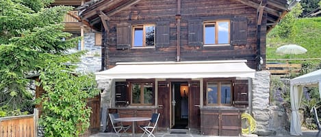 Un chalet de madera con fachada de piedra, un porche cubierto con mesa y sillas, y vistas al verde circundante.