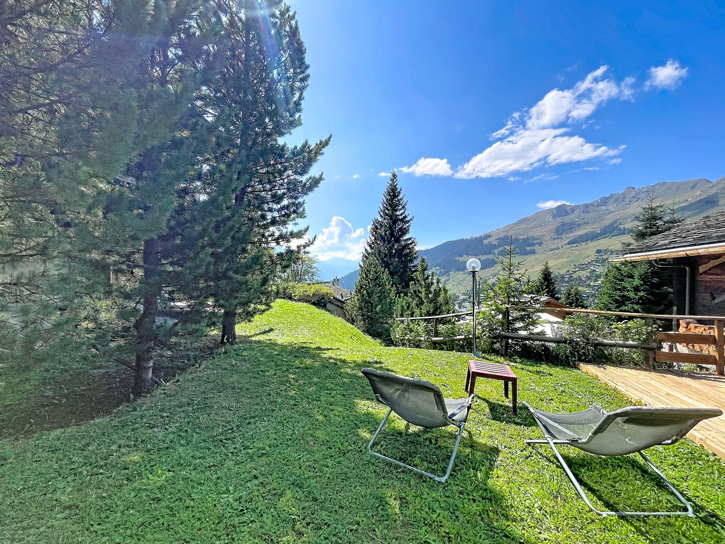 Eine grüne Außenanlage mit zwei Metallstühlen und einem kleinen Tisch, umgeben von Bäumen und Blick auf eine Berglandschaft.