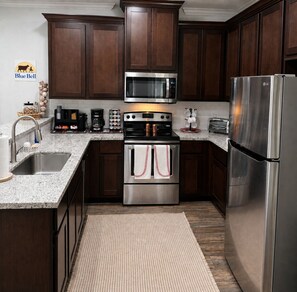 A kitchen with dark wood cabinets, stainless steel appliances including a microwave, oven, and refrigerator, a granite countertop, and a woven rug.