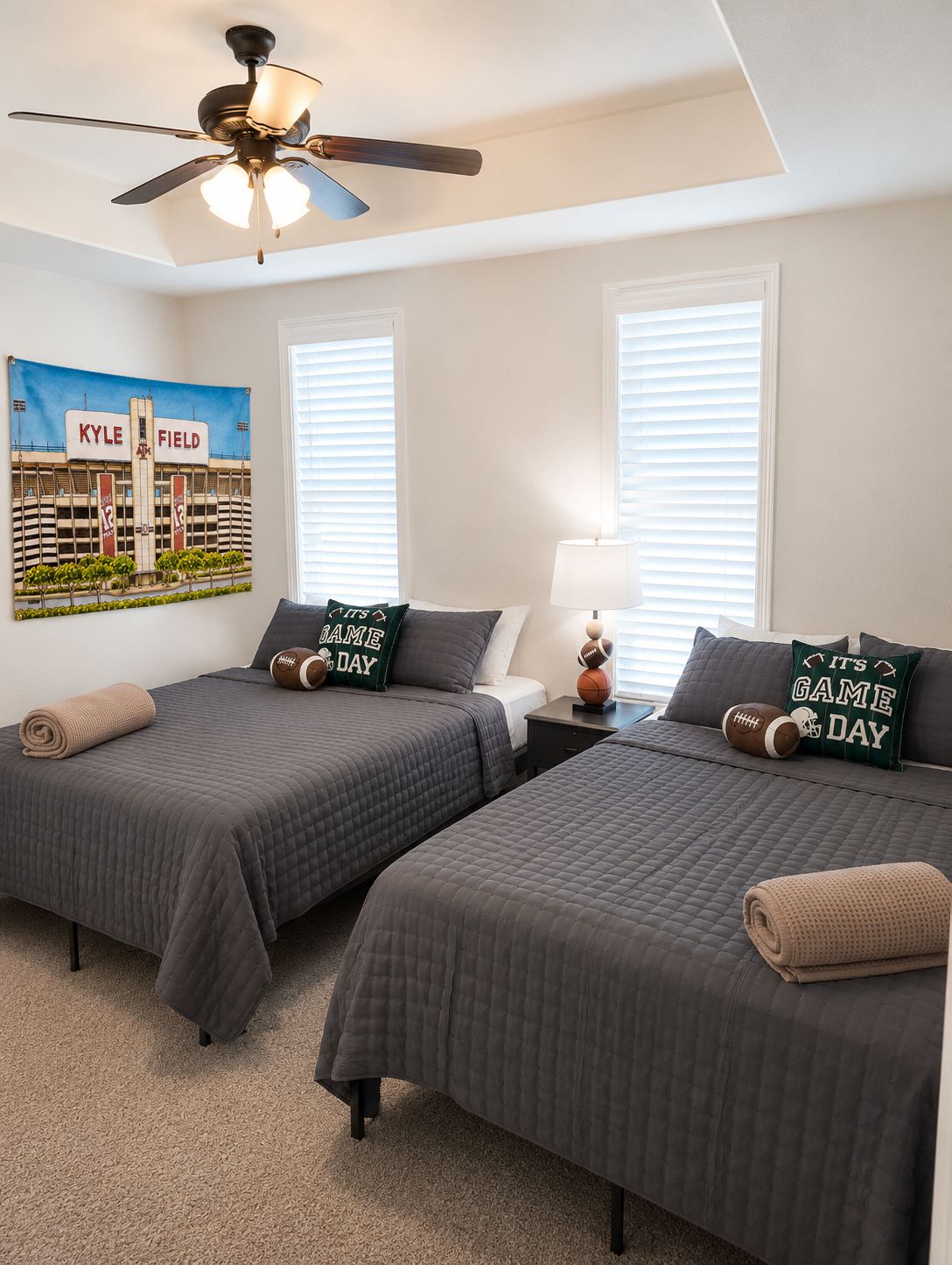 A bedroom with two beds, a ceiling fan, a lamp, and a poster of a stadium.