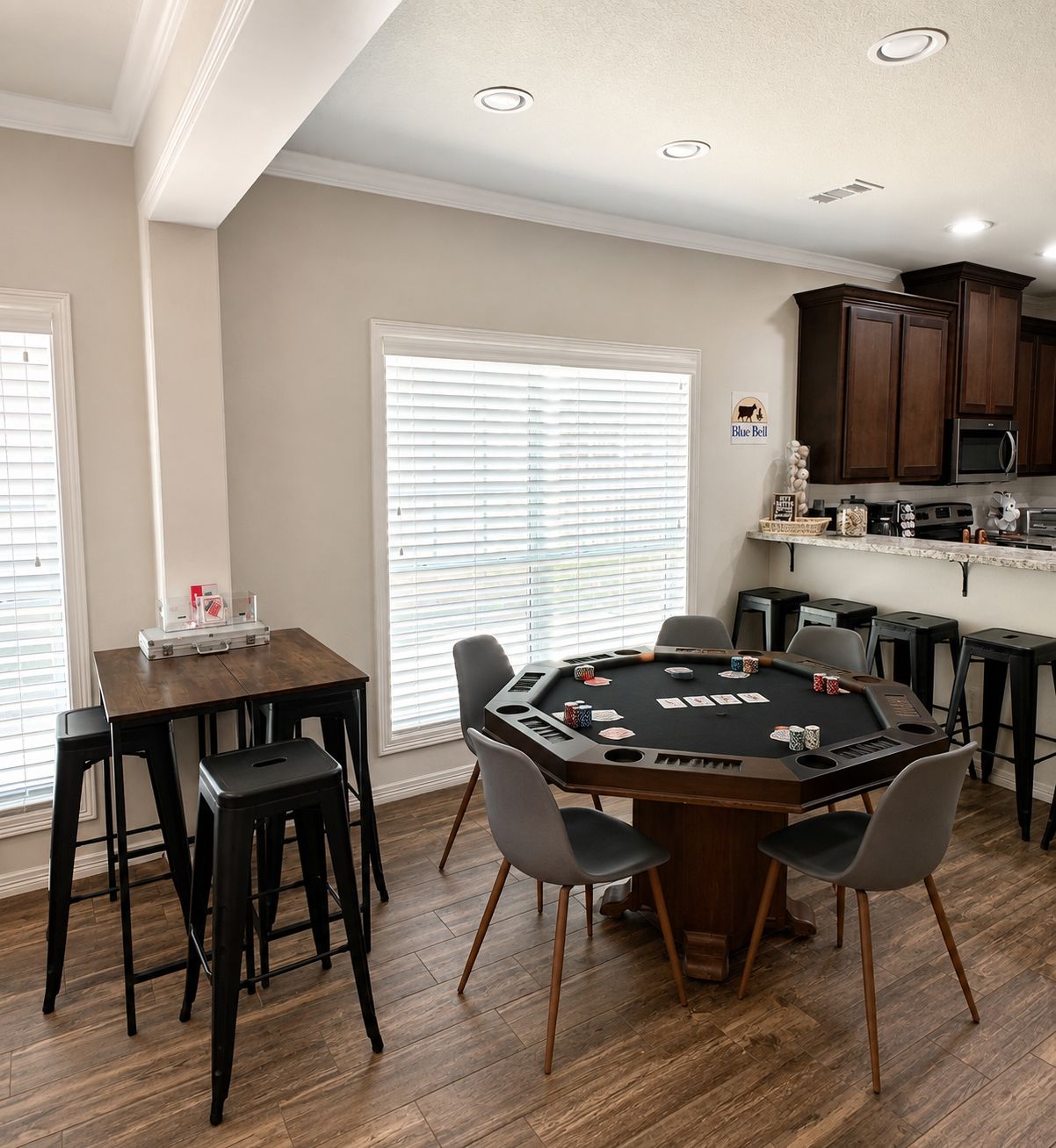 Une pièce comprenant une table de poker, des chaises, un coin cuisine et une fenêtre avec des stores.