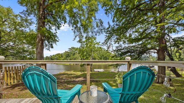Een houten terras met turquoise stoelen en een tafel, met uitzicht op een meer met een houten omheining en bomen.