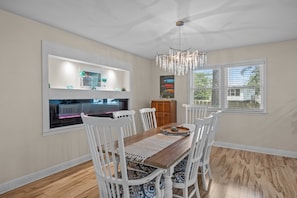 A dining area with a wooden table, white chairs, a chandelier, and a view of the outdoors through windows. - Turtle Time Groove - Waterfront Dock & Kayaks (New Bern)