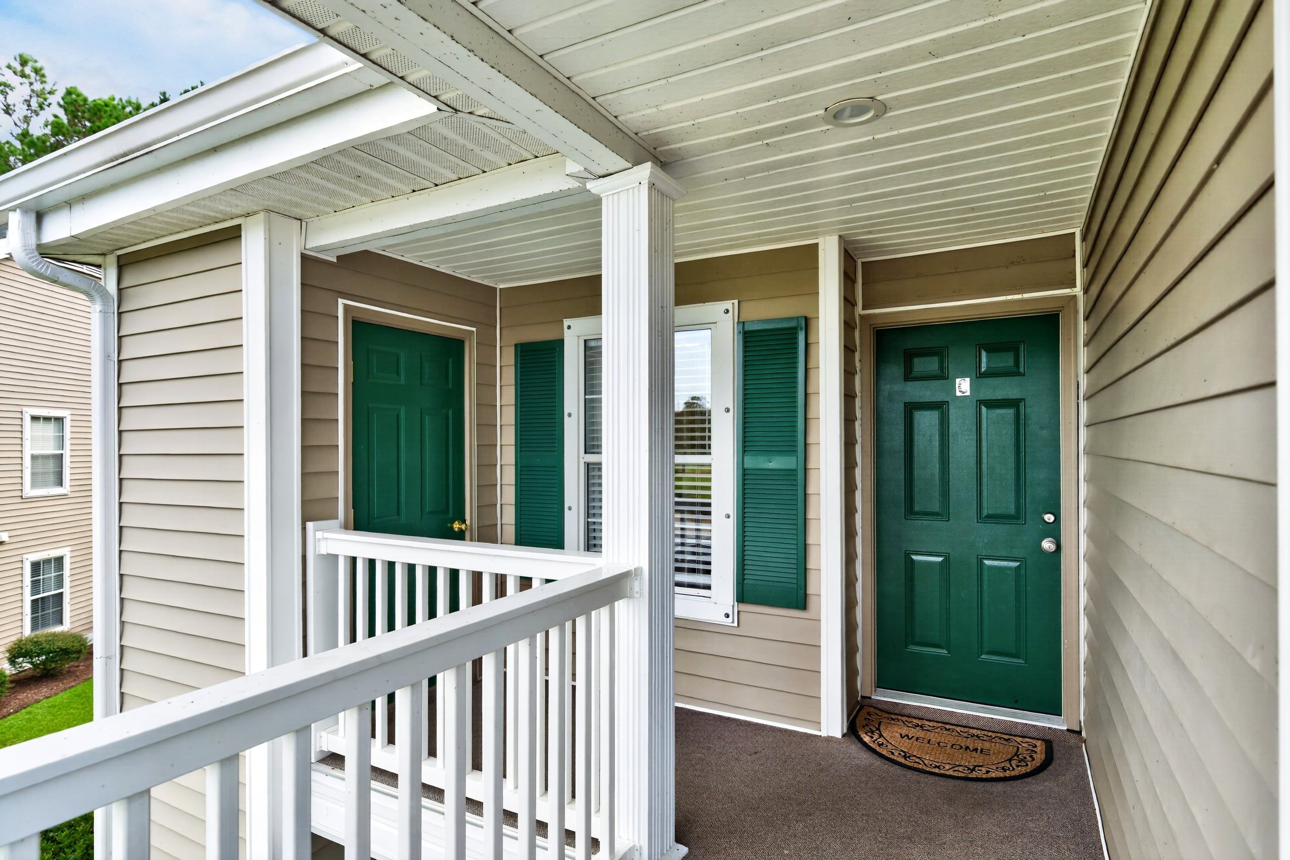 Een overdekte veranda met een witte railing, een groene deur en een welkomstmat.