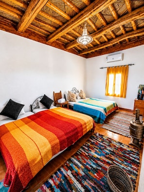 A room with two beds, a wooden ceiling, a chandelier, a window with curtains, and a colorful rug.