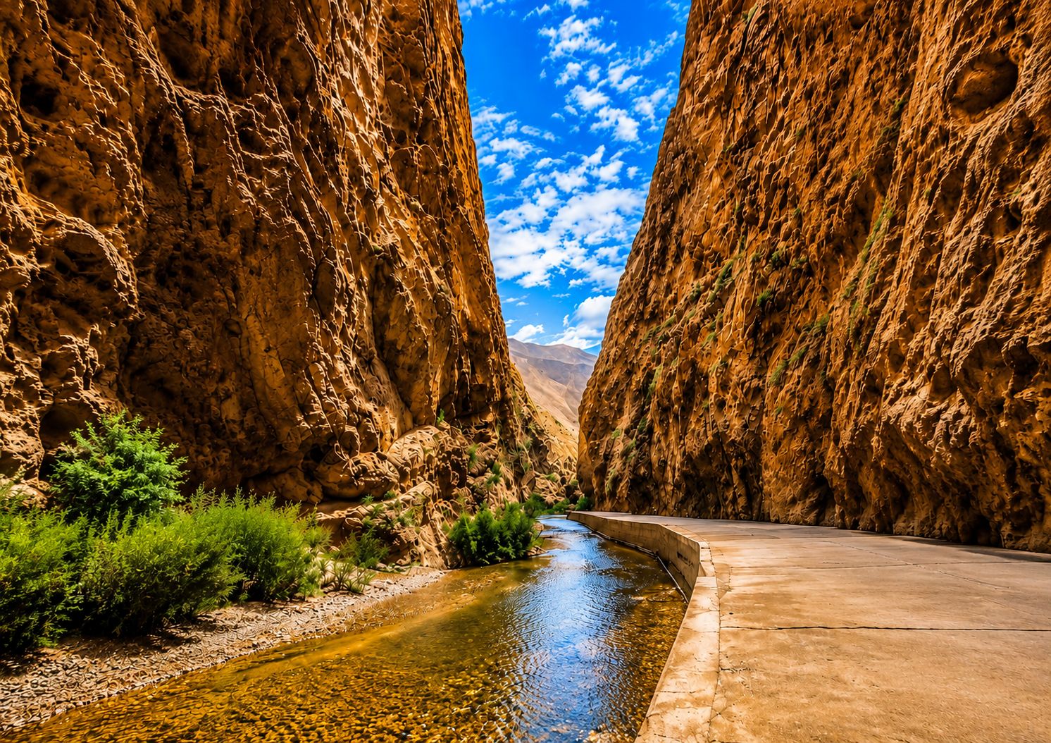 A narrow canyon with a clear stream, rocky cliffs, and a paved road.