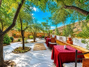 Un área de comedor al aire libre con mesas y sillas, rodeada de árboles y un muro de piedra.