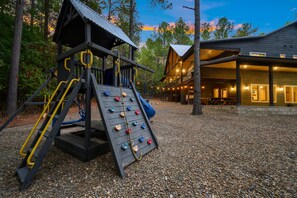 A playground with climbing and sliding equipment, a wooden building with a porch, and a gravel ground. - Luxury 5BR Cabin Near Broken Bow Lake with Hot Tub, Game Room & Playground (Broken Bow)