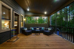 A covered outdoor seating area with a wooden deck, a sofa set, and a view of the surrounding forest. - Luxury 5BR Cabin Near Broken Bow Lake with Hot Tub, Game Room & Playground (Broken Bow)