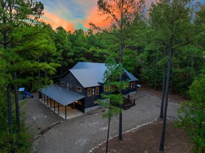 A two-story cabin with a metal roof, surrounded by dense forest, and a gravel driveway leading to the entrance. - Luxury 5BR Cabin Near Broken Bow Lake with Hot Tub, Game Room & Playground (Broken Bow)