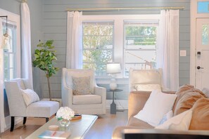 A living room with two armchairs, a sofa, a coffee table, and a lamp. There are windows with curtains, a plant, and a view of the outdoors. - NEW! The Seabreeze! Fenced Backyard, Private Parking, King Bed, Pet Friendly (Wilmington)
