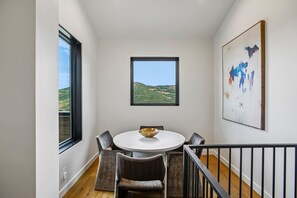 Uma sala de jantar com uma mesa redonda, cadeiras, uma tigela, uma janela com vista para uma paisagem montanhosa e um quadro abstrato emoldurado na parede.