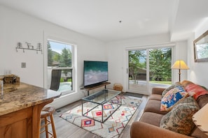 Ein modernes Wohnzimmer mit großem Flachbildfernseher, einem Glastisch, einer Couch mit gemusterten Kissen sowie einer Küchenzeile mit Marmorarbeitsplatte und Barhocker.