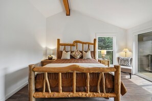 Ein hölzernes Bett mit Lattenrost, ein Nachttisch mit Lampe und Blick ins Freie durch ein Fenster.