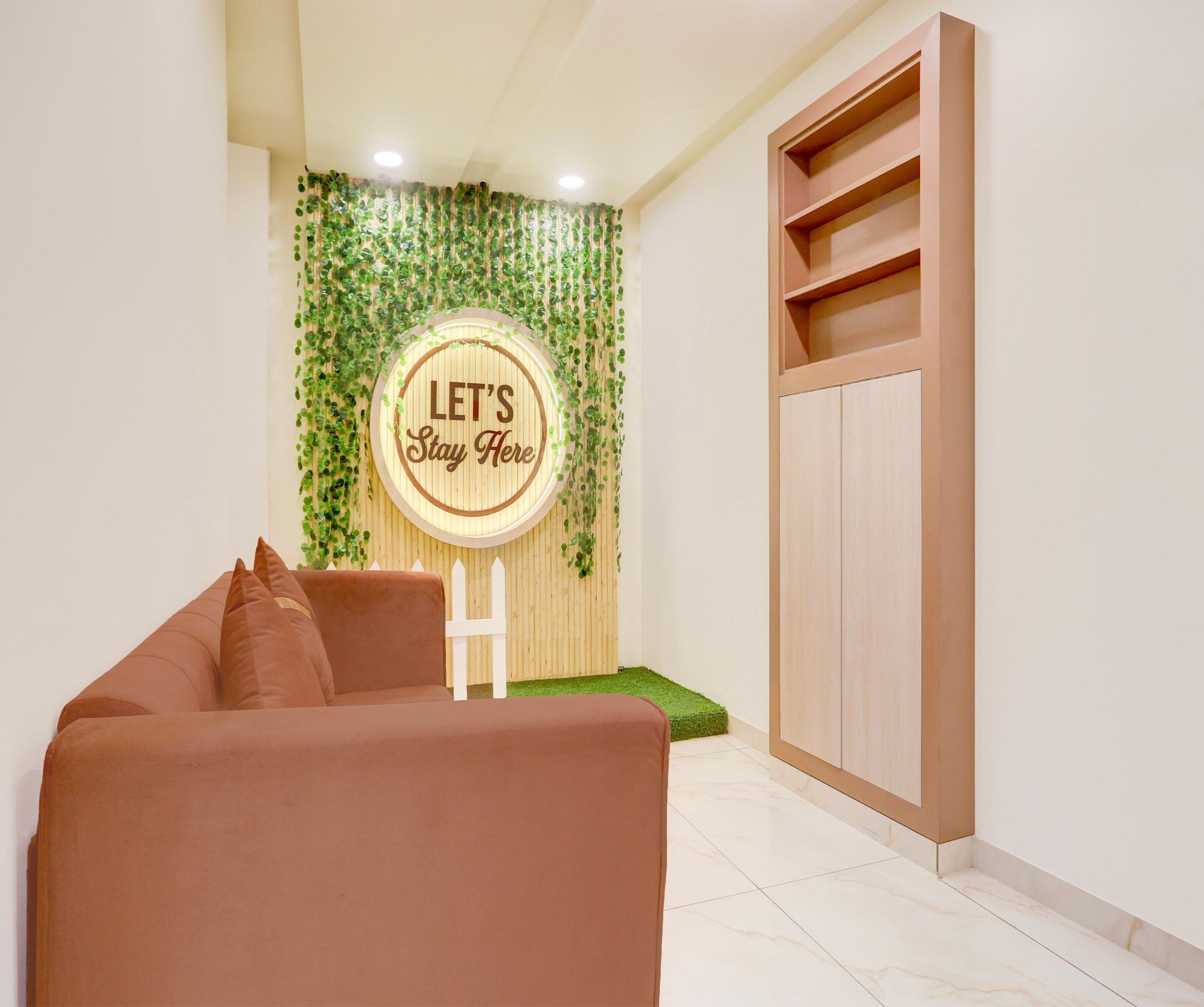 A room with a brown sofa, a wooden cabinet, and a wall decorated with green foliage and a circular sign that reads 'LET'S Stay Here'.