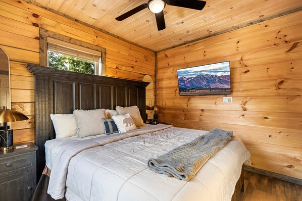 Ein Zimmer mit Holzboden, einem Bett, einem Nachttisch, einer Lampe, einem Deckenventilator und einem Fernseher an der Wand.