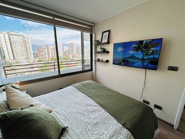 Een slaapkamer met een groot raam, een bed met beddengoed met een patroon, een bevestigde televisie en uitzicht op de stad.