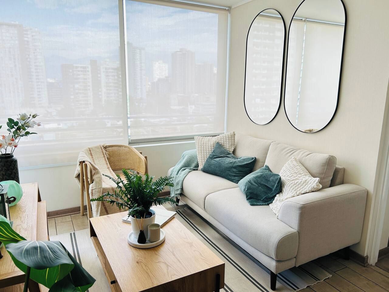 A modern living room with a sofa, a chair, a table with a plant, and a city view through large windows.