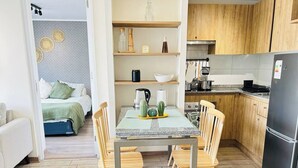 A dining area with a table and chairs, a kitchen with wooden cabinets, and a bed with a green bedspread in the background. - 2BR/2B Suite, A/C and heating, Parking (Estación Central)
