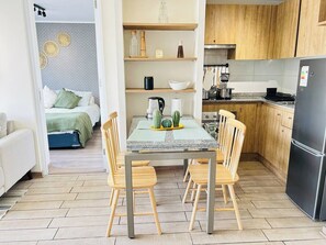 Zona pranzo con tavolo e sedie, cucina con mobili in legno e un letto con copripiumino verde sullo sfondo.