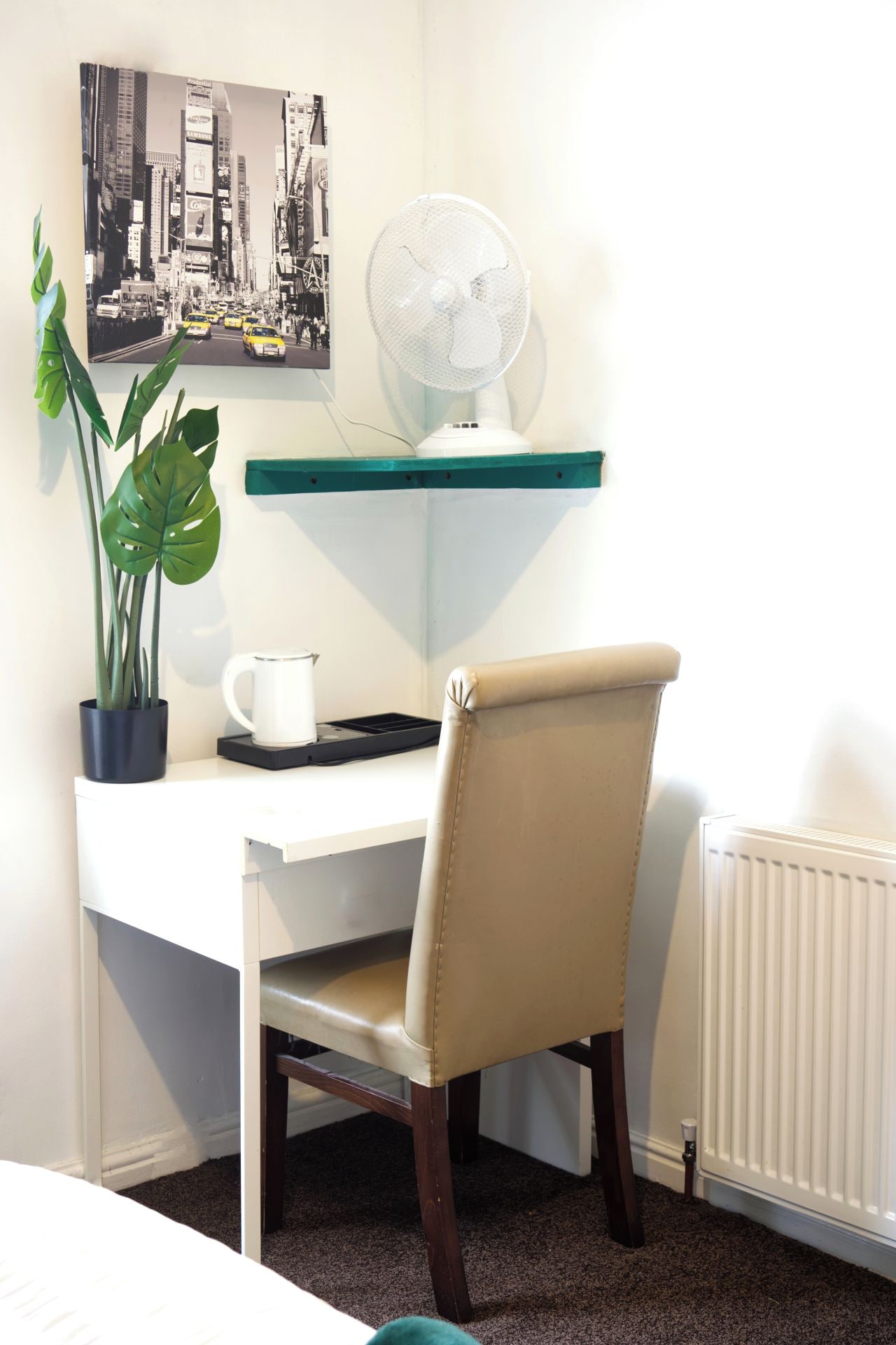 A corner office with a white desk, a chair, a potted plant, a coffee maker, a fan, and a framed cityscape picture.