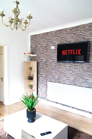 Ein modernes Wohnzimmer mit Wand im Steinoptik, einem angebrachten Fernseher, der 'NETFLIX' zeigt, einem Holzregal, einer Topfpflanze und einem Chandelier.