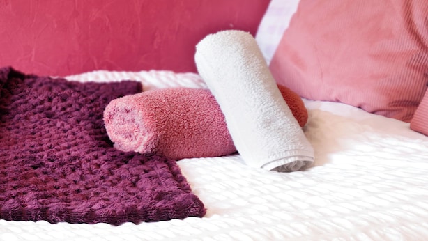 Un lit avec une couette blanche, une couverture violette tricotée et une serviette rose.