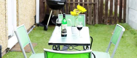 A small patio with a table and chairs, a grill, and a wooden fence in the background.