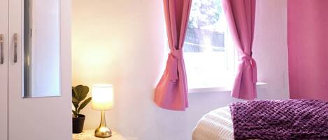 A bedroom with a bed, a desk with a lamp, a wardrobe, and pink curtains.