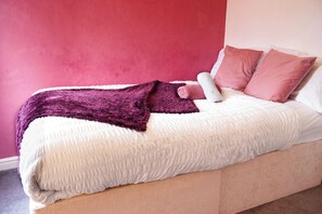 A bed with a white and purple blanket, two pink pillows, and a rolled towel.