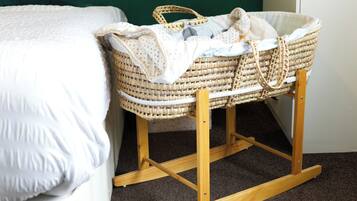A baby cradle with a woven basket seat and a wooden stand, placed next to a bed with a white quilt.