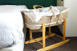Una culla per bambini con un cestino intrecciato e una struttura in legno, posizionata accanto a un letto con copripiumino bianco.