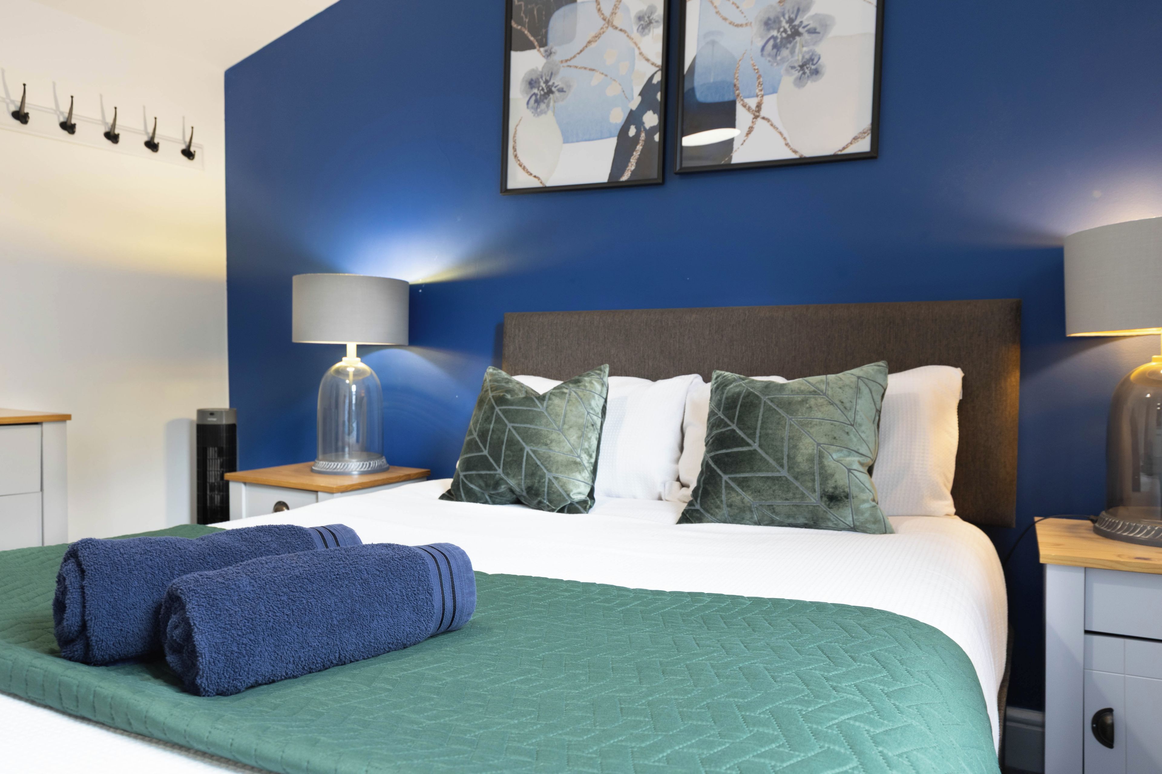 Un lit bien fait, recouvert d’une couette verte, avec deux oreillers et une serviette bleue. Une lampe est posée sur une table de chevet, et un tableau encadré orne le mur.