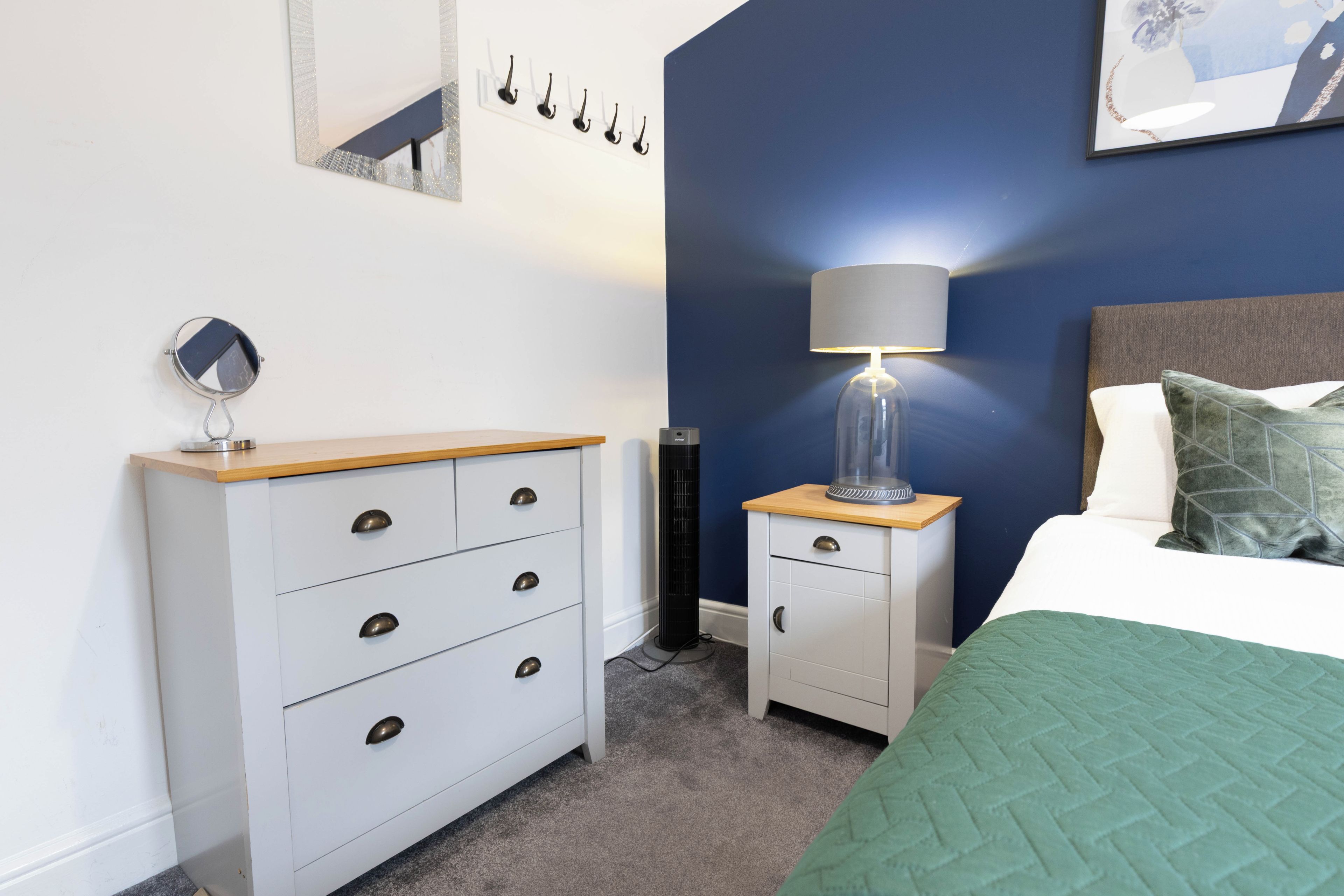 Une chambre à coucher comprenant un lit, une table de chevet, une commode, une lampe et un miroir fixé au mur.