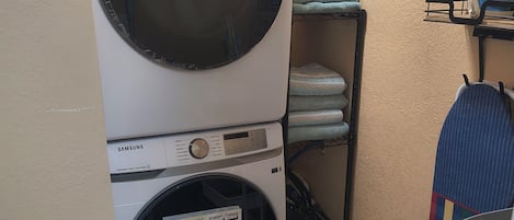 Un área de lavandería con lavadora y secadora Samsung, un estante con toallas dobladas y un tendedero con una toalla azul.