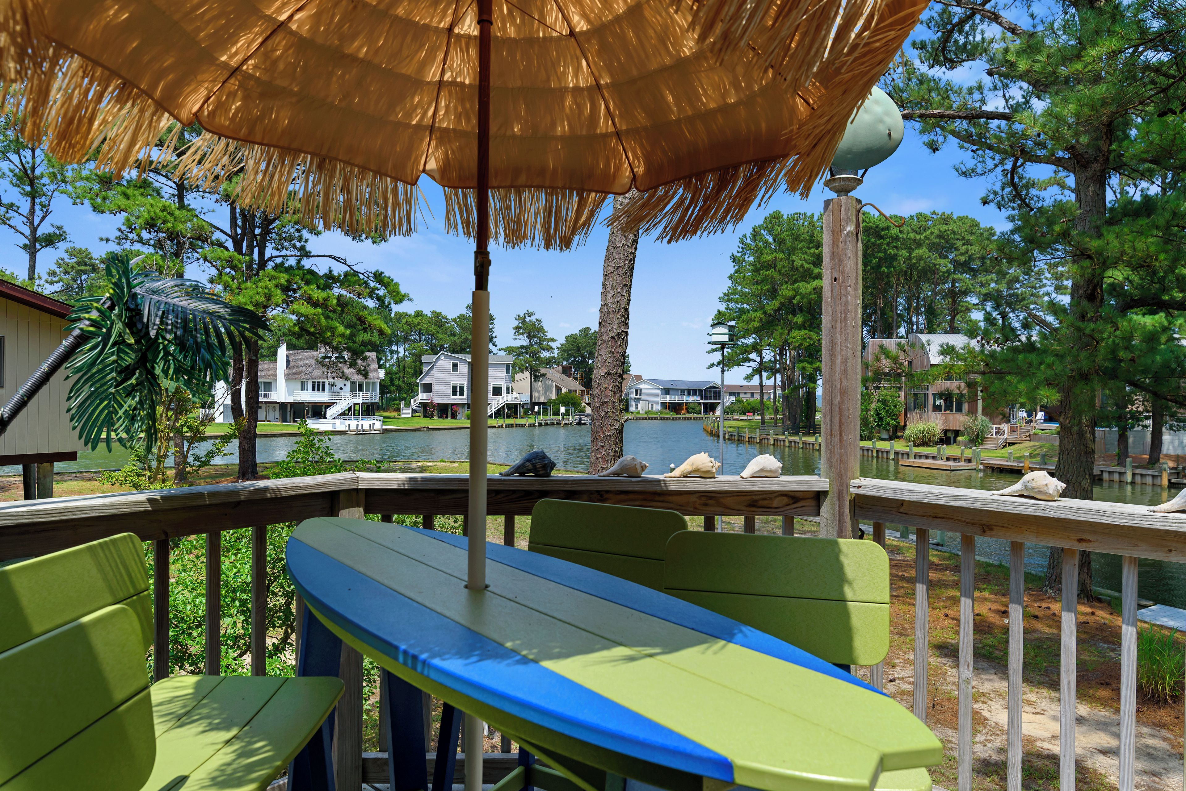 Une terrasse dotée d’un parasol en chaume, de chaises vertes et offrant une vue sur un lac, avec des maisons en arrière-plan.