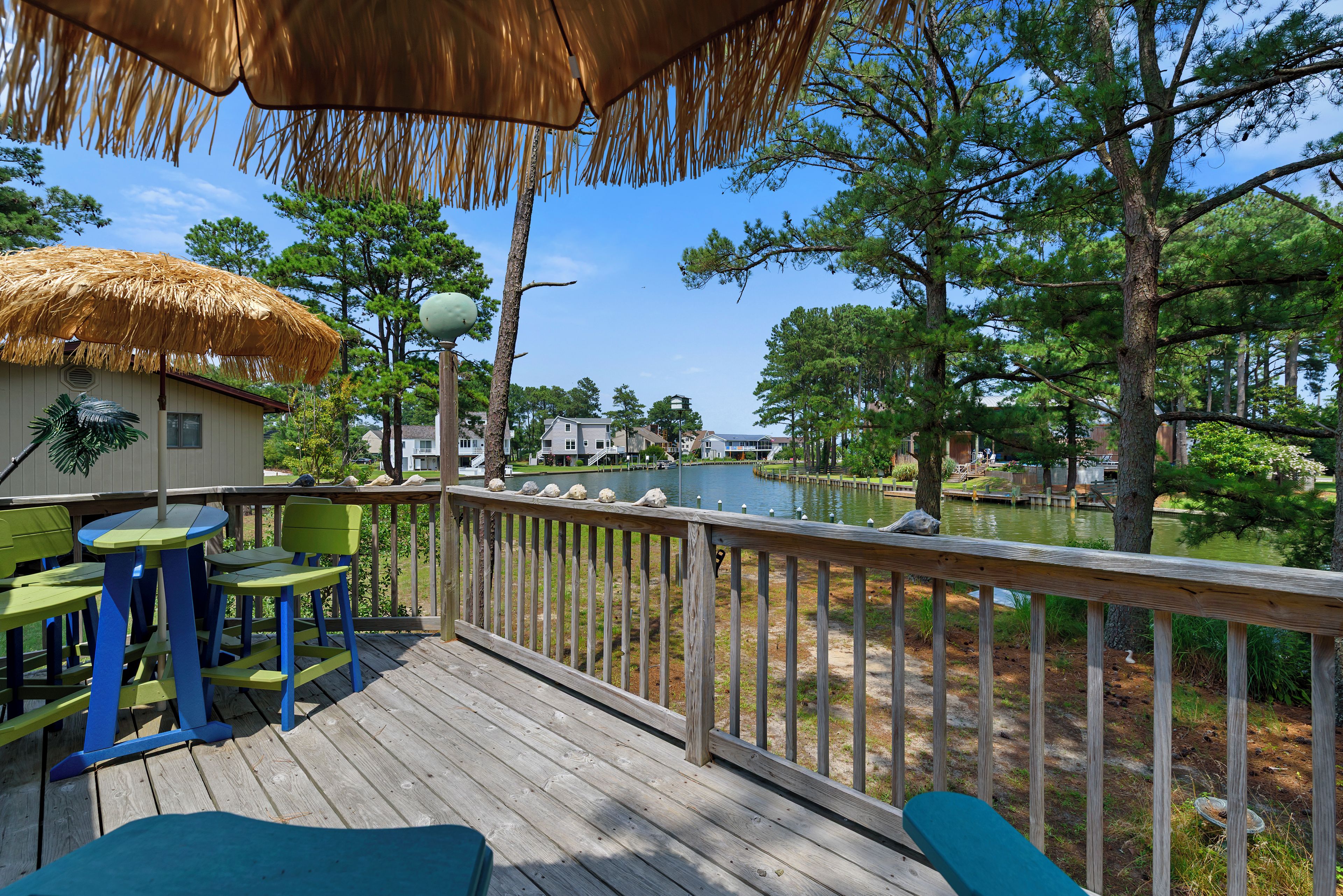 A wooden deck with a thatched roof, green chairs, and a view of a lake and buildings in the distance.