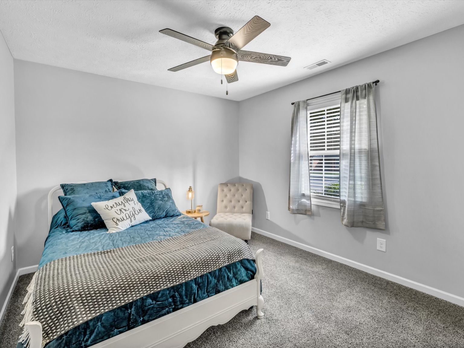 A bedroom with a bed, a chair, a ceiling fan, a window with curtains, and a nightstand with a lamp.