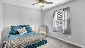 A bedroom with a bed, a chair, a ceiling fan, a window with curtains, and a nightstand with a lamp.