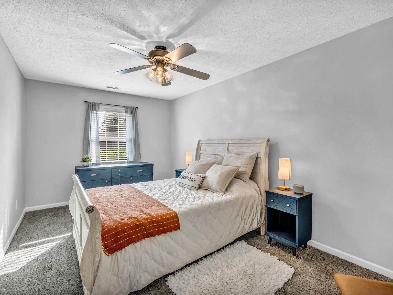 Une chambre à coucher comprenant un lit, des tables de chevet, un ventilateur de plafond, une fenêtre avec des rideaux et un tapis.