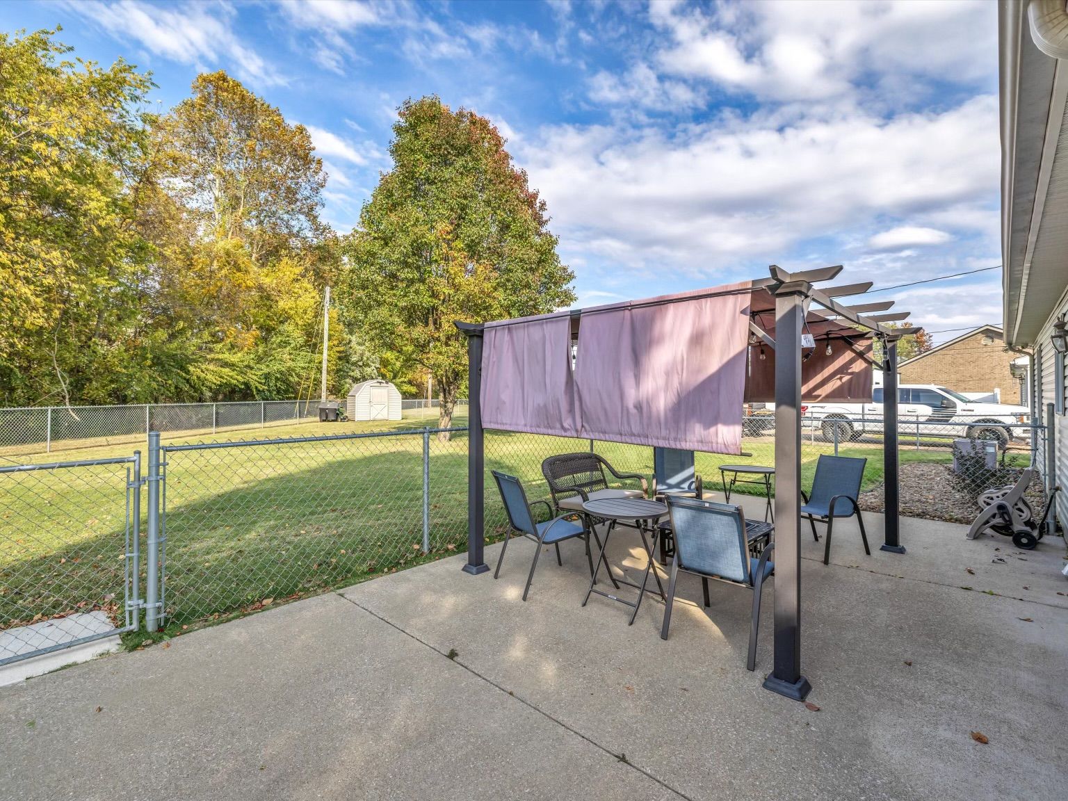 An outdoor patio area with a pergola, seating, and a view of a grassy field and trees.