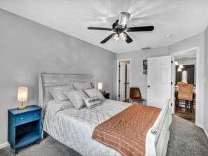 Une chambre à coucher comprenant un lit, des tables de chevet, un ventilateur de plafond et donnant sur la salle à manger.