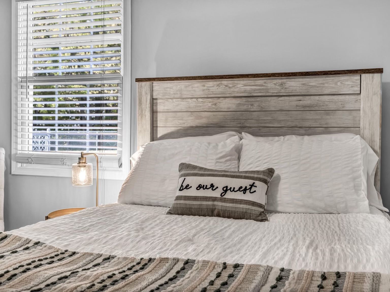 En pænt redt seng med en pude, der har teksten 'be our guest', og en sengetårnslampe.