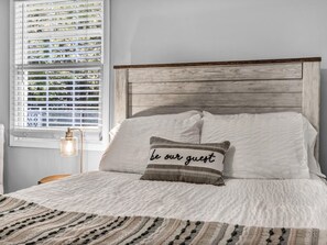 A neatly made bed with a pillow featuring the text 'be our guest' and a bedside lamp.