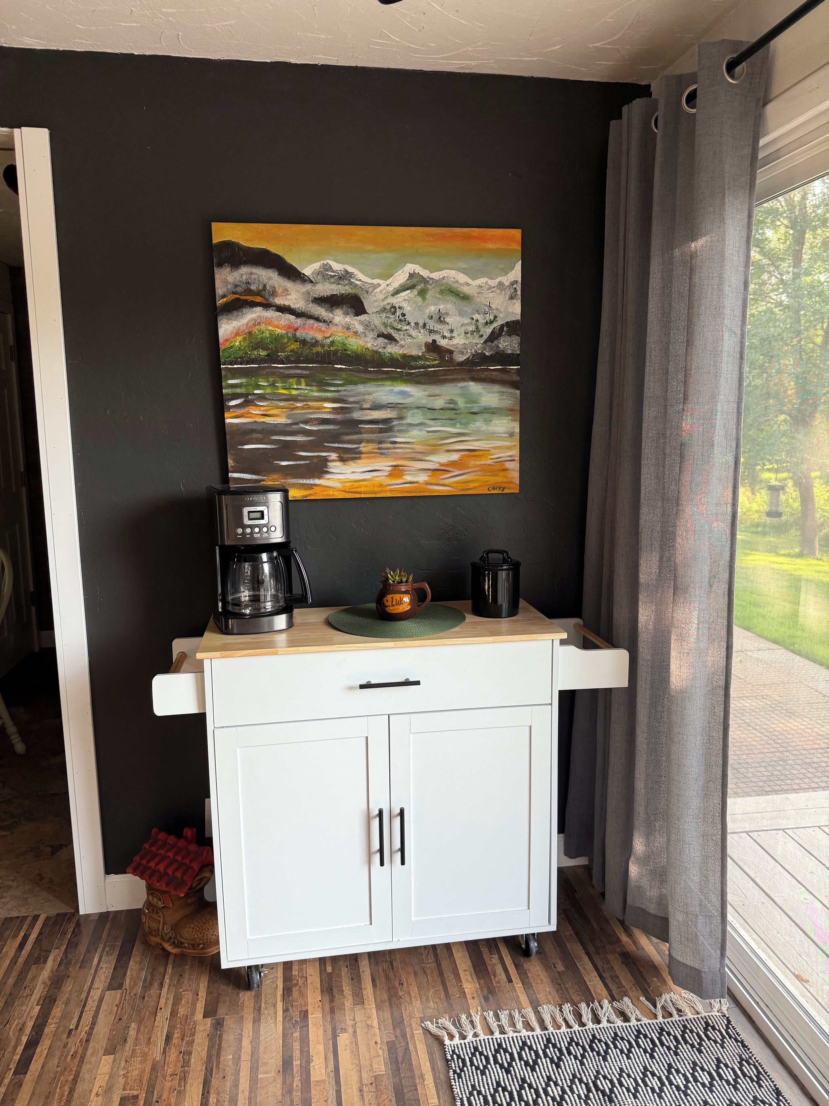A kitchenette with a coffee maker, a mug, and a painting of a mountain landscape.
