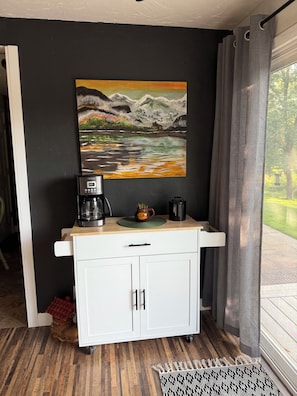 Een kitchenette met een koffiezetapparaat, een mok en een schilderij van een berglandschap.